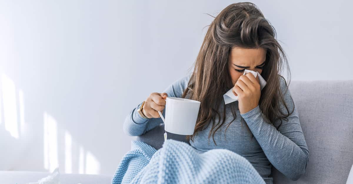 Woman blowing her nose as she holds cup of coffee or tea.