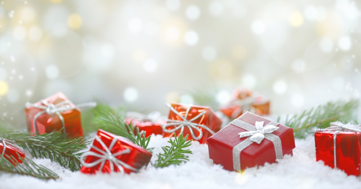 Holiday gifts wrapped in red paper with silver ribbons and bows in a holiday setting with snow, greens and lights.